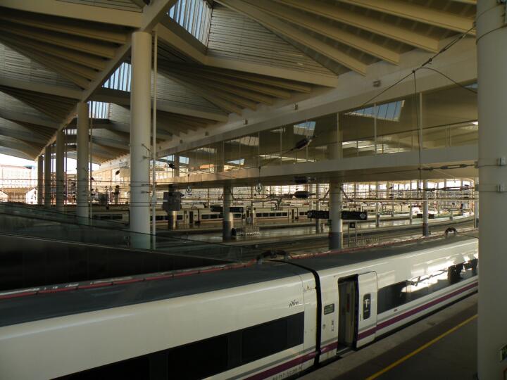 White Renfe AVE S-112 high-speed train standing at a platform inside Madrid's Puerta de Atocha station, doors open, under a steel and glass canopy