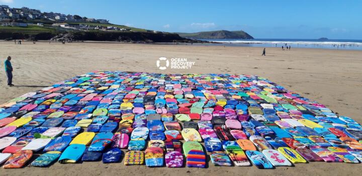 Hundreds of recovered bodyboards laid out in long rows on a beach for the Ocean Recovery Project, a person in a blue jacket standing to one side and a coastal town on the hill behind