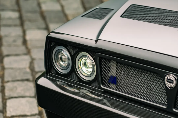 Close-up of the front of a Lancia Delta Futurista restomod: quad round headlights, a black mesh grille carrying the Lancia badge, a carbon fibre splitter and a silver-grey bonnet over cobblestones