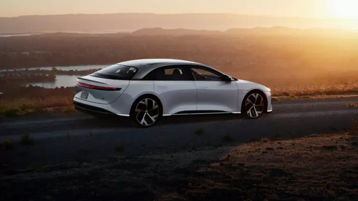 A white Lucid Air sedan on a winding road at sunset, overlooking a valley with a river and hazy hills in the distance